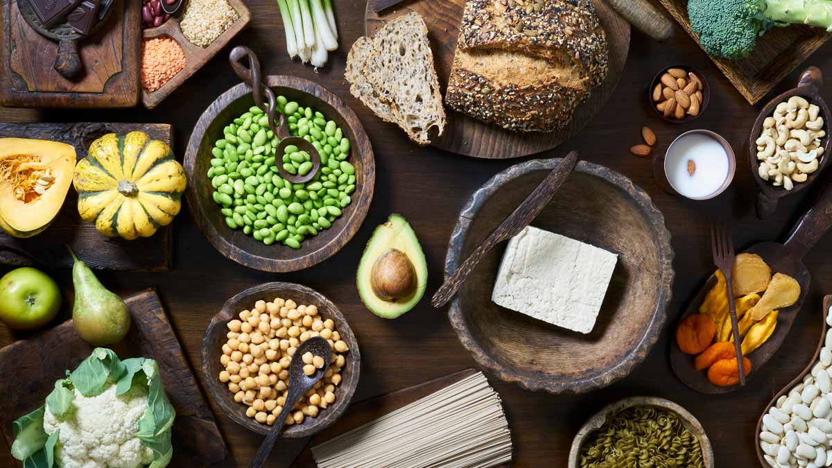 overhead view of wooden bowls and boards with beans, nuts, tofu, whole grain bread, squash, broccoli, cauliflower, avocado, and dried fruit
