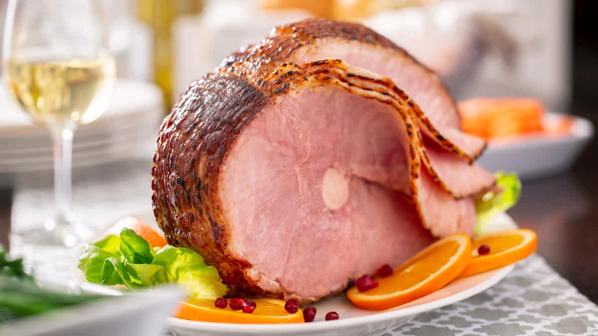 close up of bone-in spiral ham on white plate with pomegranate seeds, lettuce, and orange slices and glass of wine and stack of plates blurred in background