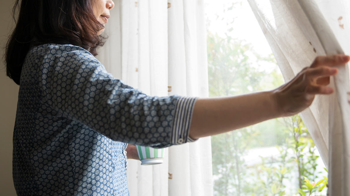 Woman pulling back curtains holding a cup looking outside