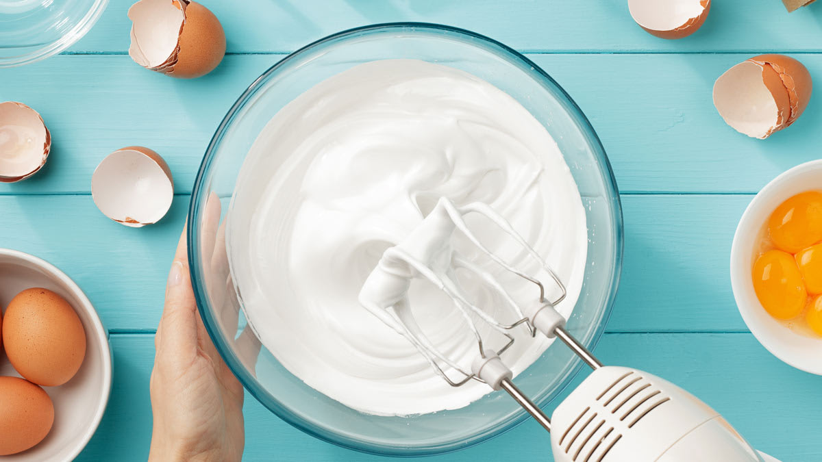Hand using an electric mixer to whip egg whites in a glass bowl, surrounded by cracked eggshells, whole eggs, egg yolks in a small bowl, and baking tools on a turquoise wooden surface.
