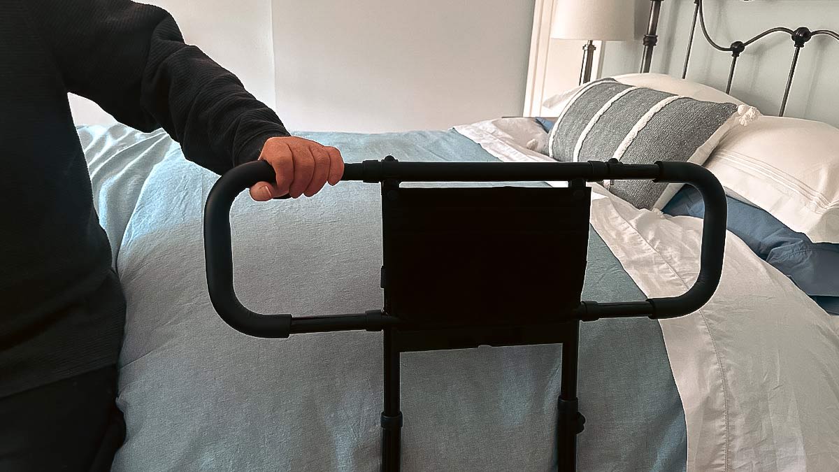 A person's hand grips a black, U-shaped metal bed assist rail that is positioned against the side of a bed in a residential bedroom.