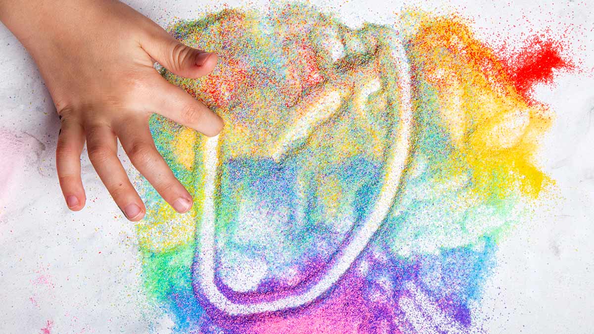 Child's hand creating a rainbow shape in a mixture of colorful sand.