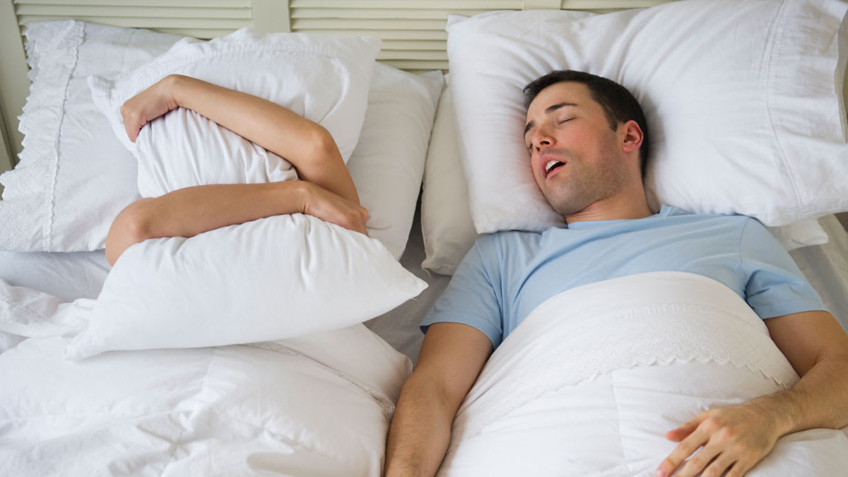 Couple in bed with man snoring and woman covering face with pillow