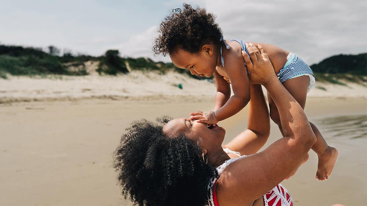A smiling mother playfully lifts her toddler into the air on a beach with sand dunes in the background.