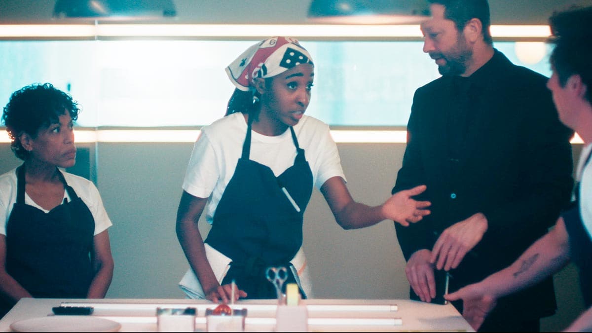 A tense kitchen scene from The Bear featuring Liza Colón-Zayas, Ayo Edebiri, Ebon Moss-Bachrach, and Jeremy Allen White. Ayo Edebiri's character gestures emphatically while speaking, as the others look on closely in a modern, restaurant-kitchen interior.