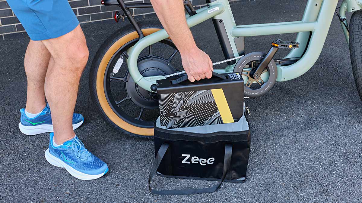 A person is shown placing a large, patterned e-bike battery into a black "Zeee" brand bag on the ground next to a mint green electric bicycle.