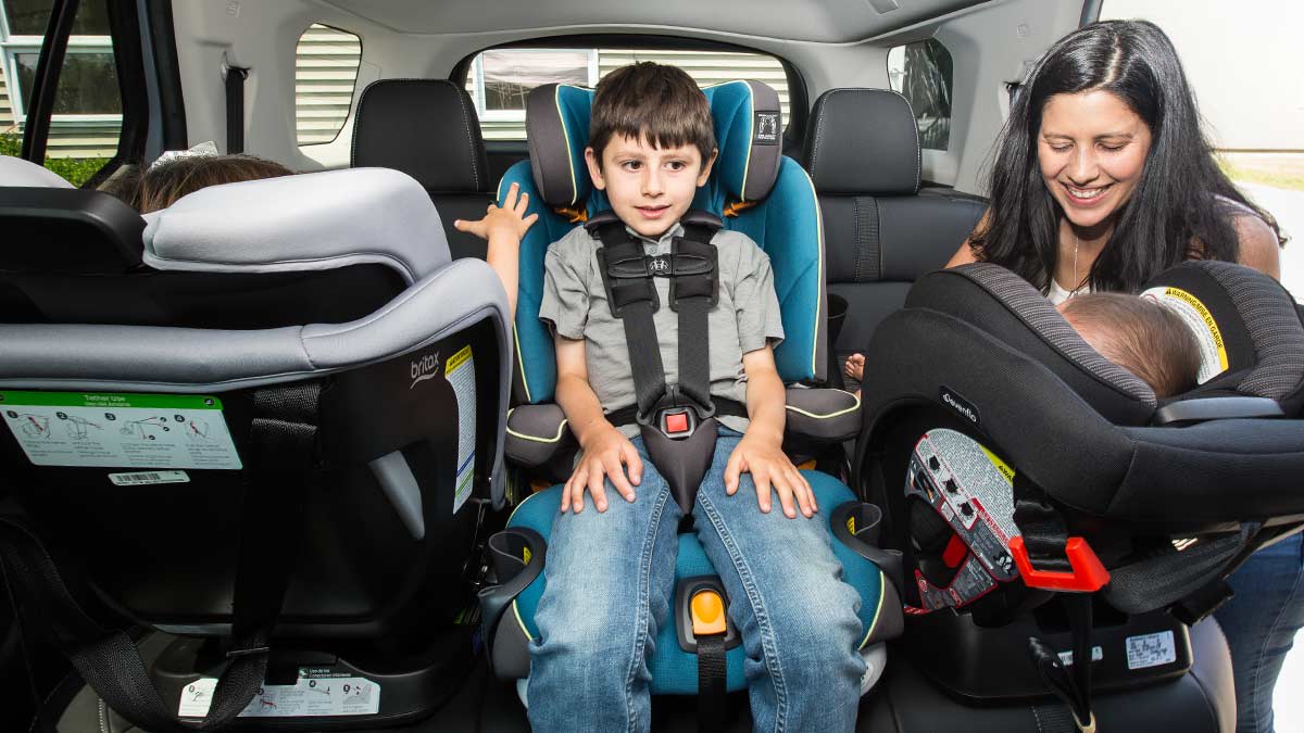 A woman smiles down at an infant in a rear-facing car seat on the right side of a vehicle's back row. In the middle, a young boy is buckled into a forward-facing teal car seat, while another child sits in a rear-facing seat on the left. This setup demonstrates a three-across car seat configuration designed to fit multiple children safely in a single row.