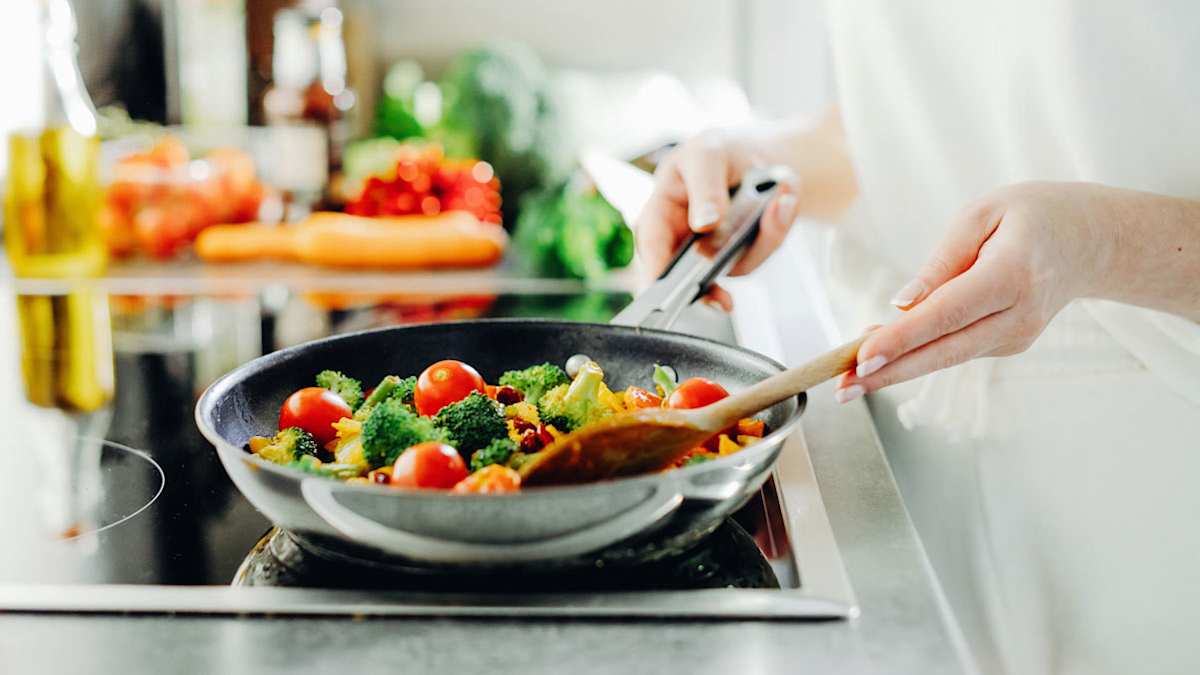 A person stirs a skillet filled with broccoli, cherry tomatoes, and other vegetables on a stovetop
