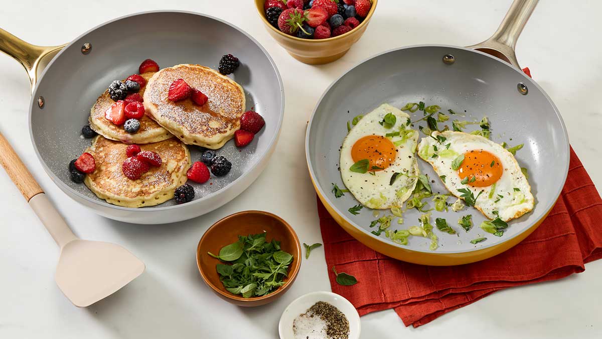 Two ceramic frying pans, one containing berry pancakes and the other two fried eggs, sit on a kitchen counter.