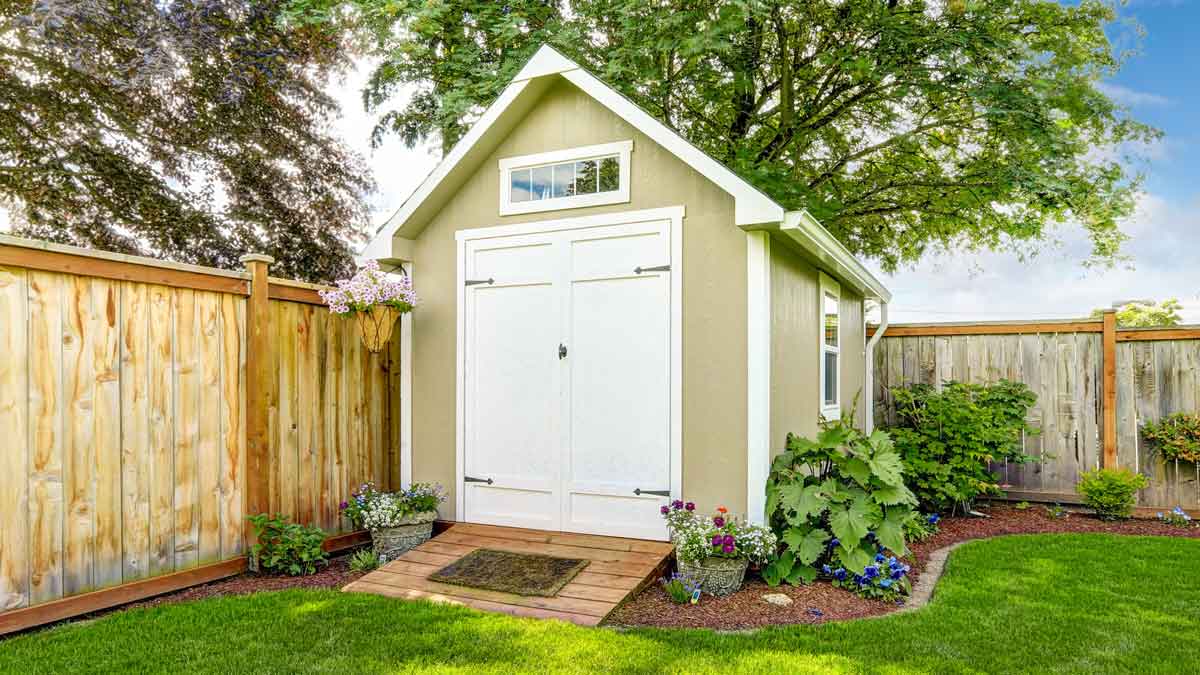 beige storage shed with white door and trim in corner or yard with wooden fencing, lawn, and plants around the shed