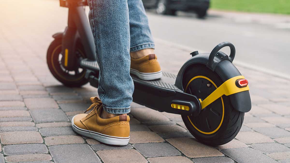 A close-up shot of a person's lower legs and feet while riding a black electric scooter on a paved cobblestone sidewalk.