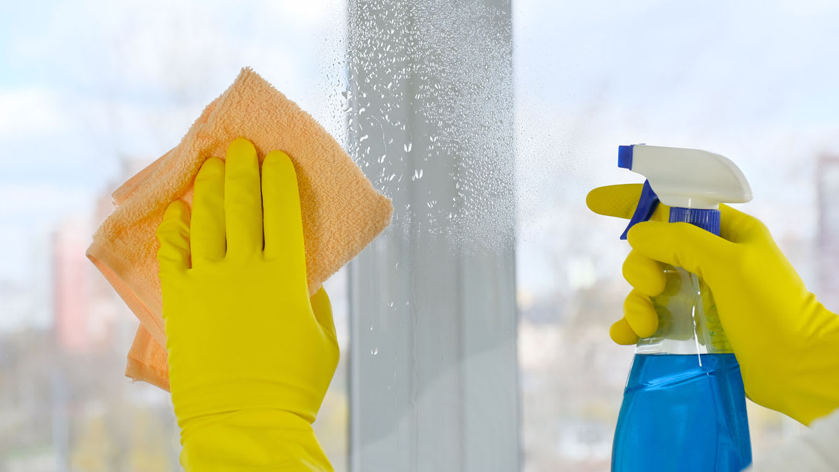 close up of hands wearing yellow latex gloves using orange microfiber cloth and glass cleaning solution to clean window
