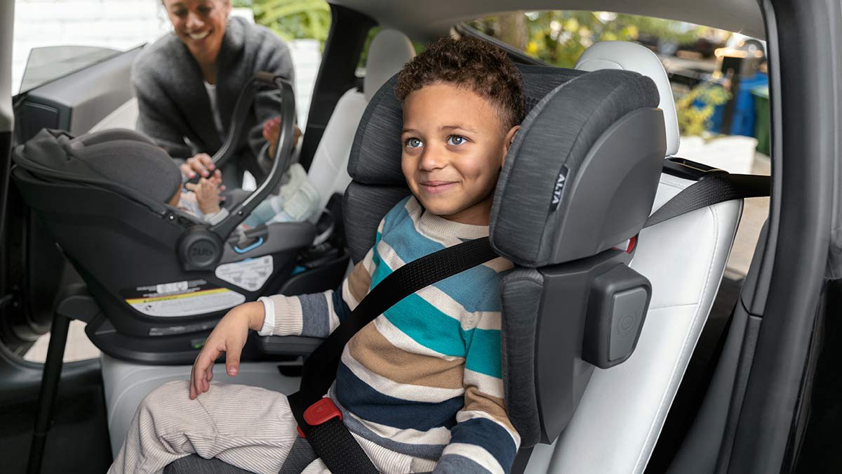 A young boy sits happily in a dark grey high-back booster seat secured by a seatbelt in the back of a car. In the background, a smiling mother leans into the open car door next to an infant resting in a rear-facing baby carrier.