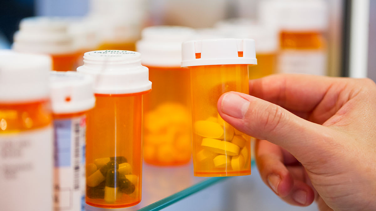 A hand taking a bottle of prescription pills out of a bathroom cabinet filled with other prescription pill bottles.