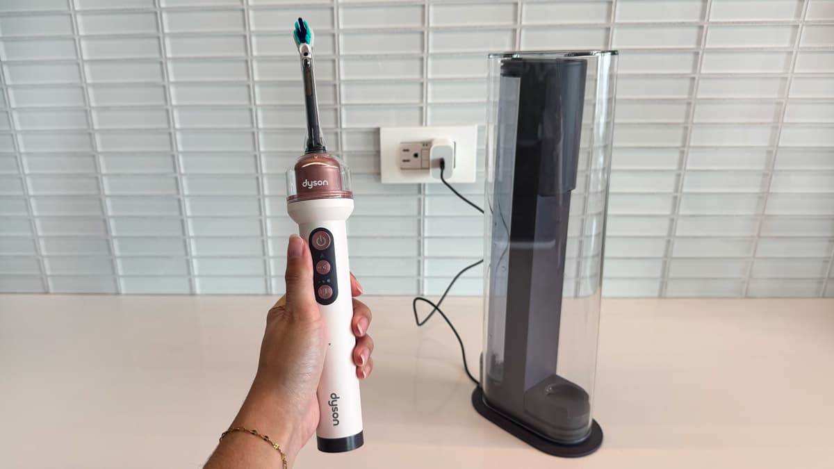 Hand holding Dyson Camerajet toothbrush next to the charging case plugged into a wall