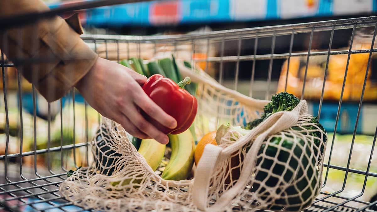 Person placing a red pepper in their shopping cart along with bananas, an orange and a head of broccoli.