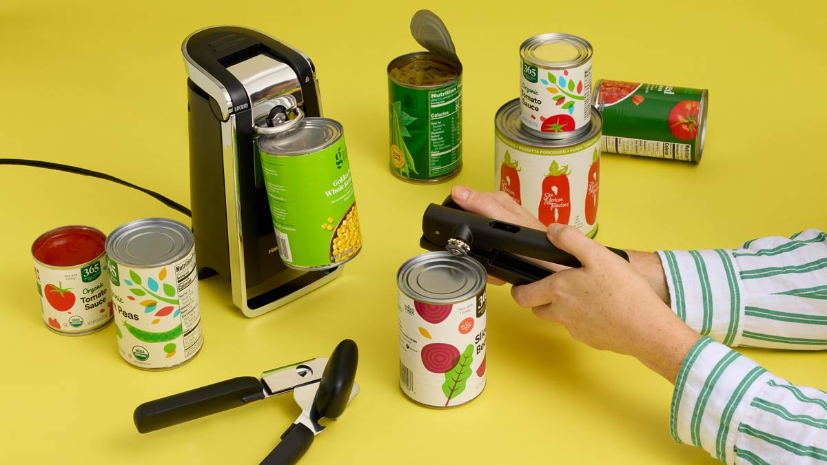 Several cans of vegetables placed near a Hamilton Beach can opener opening a can while a pair of hands uses a different can opener on a can