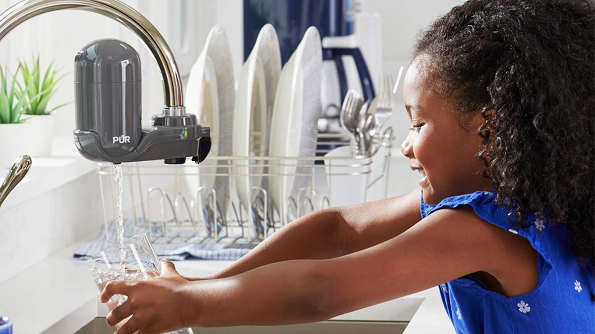 A young girl wearing a blue patterned top smiles as she fills a glass with water from a kitchen faucet attached to a gray PUR filter.