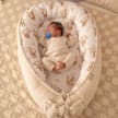 A baby seen from above asleep on their back in a CocoMi Premium Baby Nest with a pacifier in their mouth.