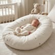 A baby asleep on their back in a Little Manta Soft Nest Baby Lounger thats sitting on top of a bed in a nursery setting.