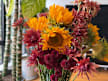 A flower arrangement featuring sunflowers, spiky reddish-orange spider mums, and deep burgundy pom-pom chrysanthemums indoors.