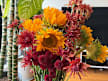 A flower arrangement featuring sunflowers, spiky reddish-orange spider mums, and deep burgundy pom-pom chrysanthemums indoors.