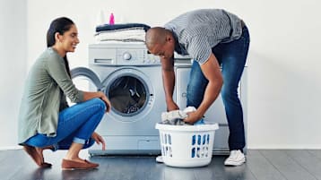 couple doing laundry in front of washer and dryer, taking laundry from basket