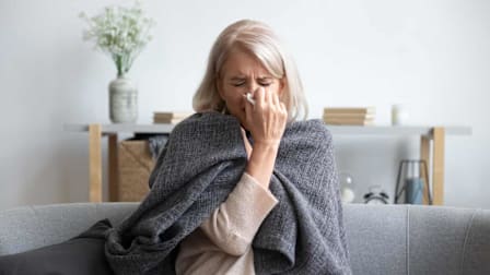 person sitting on couch wrapped in blanket and blowing their nose