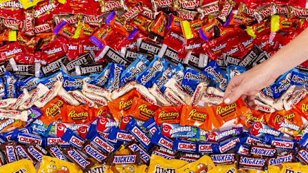 rows of various halloween candy (Twix, Reese's, Twizzlers, Hershey's Milk Chocolate, Almond Joy, Whoppers, Airheads, Crunch, Snickers, and Peanut M&Ms) with hand reaching for a Reese's Peanut Butter Cup
