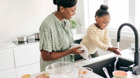 Child washing hands at kitchen sink with woman standing next to her.