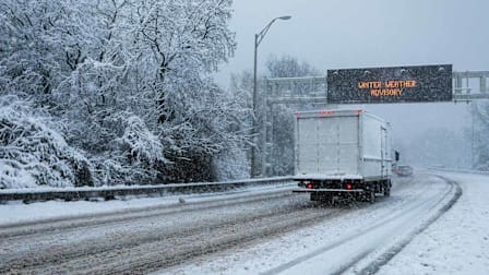 A truck driving on a highway during a winter storm with a illuminated highway sign overhead advising of a Winer Weather Advisory.