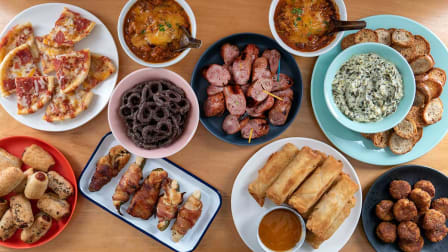 overhead view of spread of plated Superbowl snacks from Costco like spring rolls, chocolate pretzels, jalapeno poppers, chili, pizza, and spinach artichoke dip