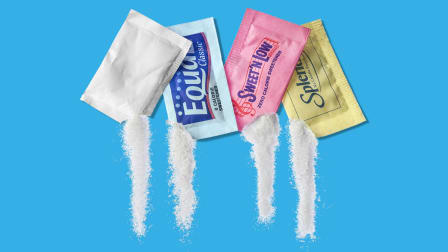 4 different artificial sugar packets with corners ripped off and artificial sugar spilling out