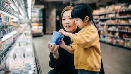 adult showing container of greek yogurt to child in grocery store aisle