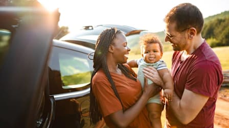 couple smiling while holding baby with car and hills in background