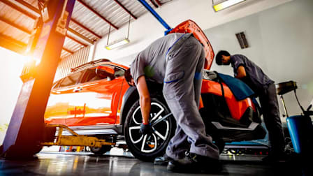 An auto repair shop with two mechanics working on an orange car.