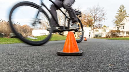 low angle view of person riding electric bike on asphalt path, going between orange traffic cones with house and grass in background
