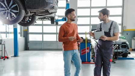 customer talking to mechanic holding tablet in auto repair shop with cars in background