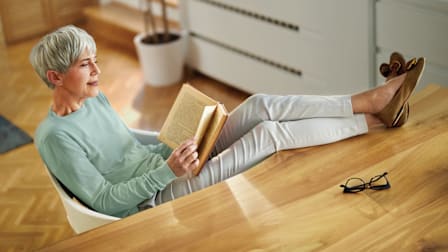 person sitting at desk with legs elevated while reading a book, glasses on desk and potted plant in background