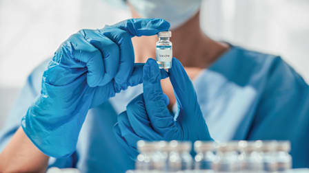 close up of person's hands holding glass vaccine vile, wearing blue surgical gloves and scrubs