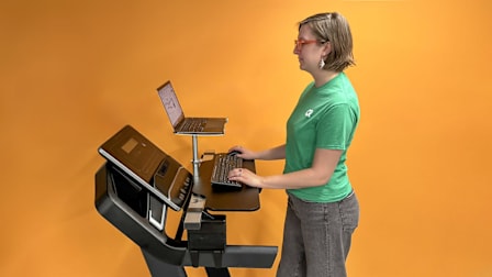 person wearing green CR branded t-shirt using laptop while standing on treadmill with RiseEight treadmill desk attachment in place