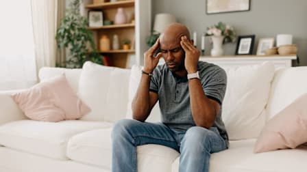 person sitting on white couch in living room with plants and shelves behind them, their eyes closed and hands on their temples as though they're in pain