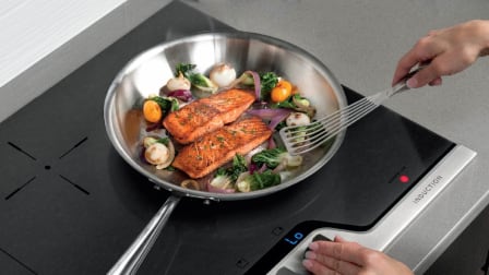 Person cooking salmon and vegetables in a stainless steel pan on an induction cooktop.