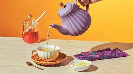 Tea being poured into a tea cup with honey and lemon slices on the table.