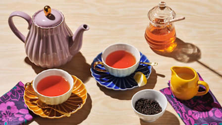A teapot next to two cups of tea, honey, milk, and loose tea leaves in a bowl.