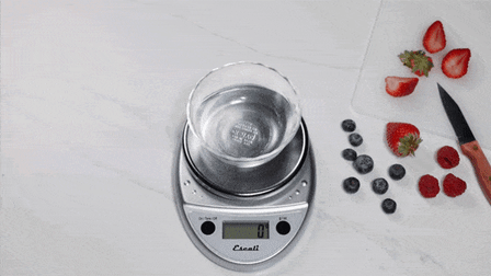 animated gif of Escali scale with glass bowl and reaching in and pouring mixed berries into bowl on scale, cutting board with mixed berries and knife to the side