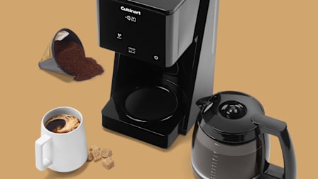 The Cuisinart DCC-T20 Touchscreen 14-Cup Programmable coffee maker on a beige background, surrounded by a reusable filter filled with spilled ground coffee. a white mug filled with swirling coffee and a few brown sugar cubes.