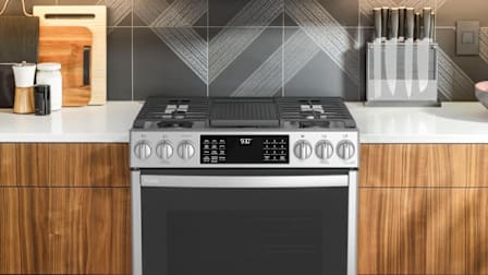 Profile PGS930YPFS range in kitchen with wooden cabinets and grey stone backsplash with knife block and cutting boards on countertop