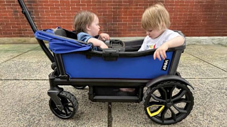 two toddlers in blue Veer baby stroller wagon on concrete sidewalk with brick wall behind them