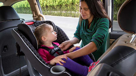 A mother buckling her toddler into a rear-facing car seat.
